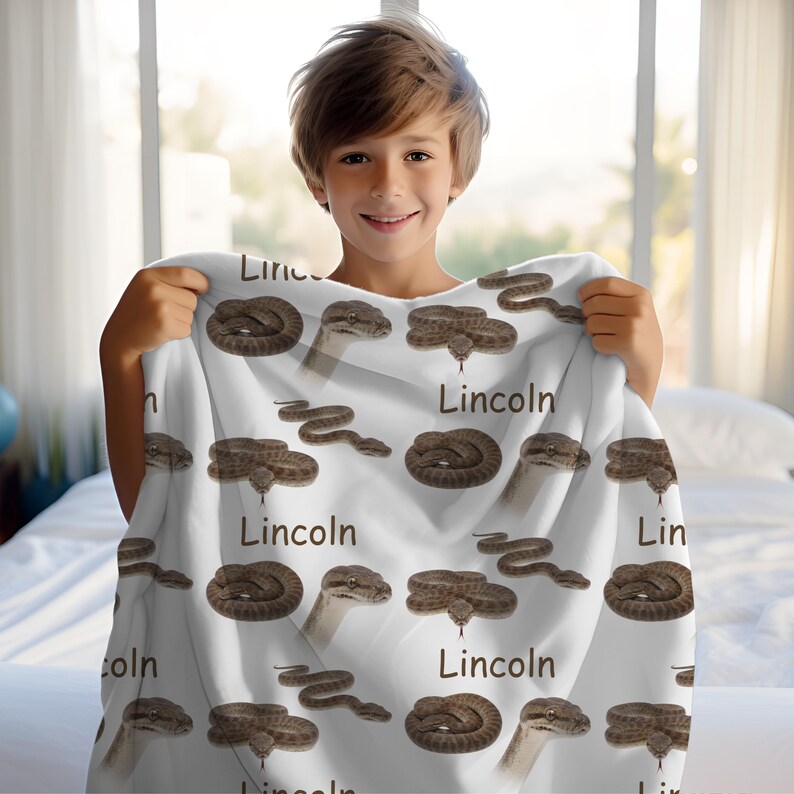 May include: White blanket featuring a repeating pattern of brown snakes and the name "Lincoln" in a stylized font. The snakes are depicted in various poses, including coiled and stretched out. The blanket is held up by a person.