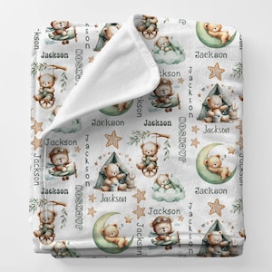 May include: White baby blanket with a repeating pattern of teddy bears in various scenes, including airplanes, swings, and tents. The name "Jackson" is also printed on the blanket, along with stars and moons.