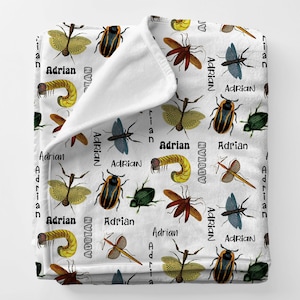 May include: White blanket featuring a pattern of various insects in shades of brown, green, and blue. The name "Adrian" is printed repeatedly in black. The blanket is folded over, revealing a soft, white interior.