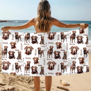 Puede incluir: Una toalla de playa blanca con un patr&oacute;n repetido de retratos e im&aacute;genes de cuerpo completo de perros Labrador Retriever marrones. El nombre "Maisy" est&aacute; impreso en escritura marr&oacute;n. La toalla se sostiene en una playa con el oc&eacute;ano al fondo.