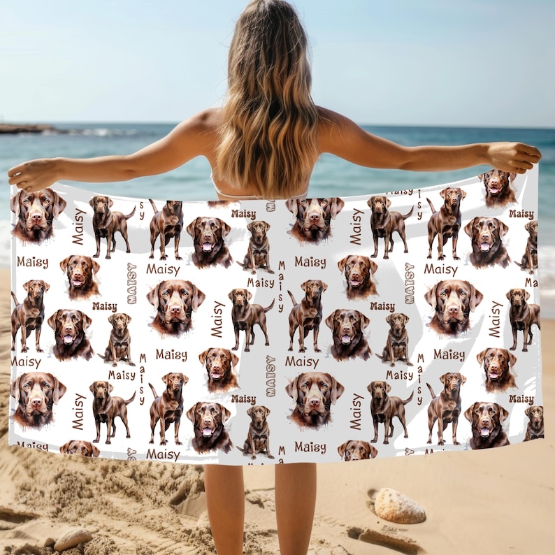 Puede incluir: Una toalla de playa blanca con un patr&oacute;n repetido de retratos e im&aacute;genes de cuerpo completo de perros Labrador Retriever marrones. El nombre "Maisy" est&aacute; impreso en escritura marr&oacute;n. La toalla se sostiene en una playa con el oc&eacute;ano al fondo.