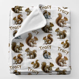 May include: White plush blanket featuring a repeating pattern of squirrels in various poses. The word "Tracy" is printed in brown script throughout the design. The blanket is folded over, revealing a white interior.