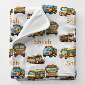 May include: White blanket with a repeating pattern of yellow school buses. The text "Shiloh" and "iloh" are printed on the blanket. The school buses are in various poses and styles.