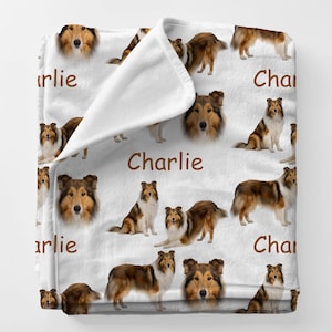 May include: White blanket featuring a repeating pattern of a Collie dog in various poses and headshots. The name "Charlie" is printed in brown script across the blanket. The dog's fur is brown, white, and tan.