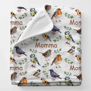 May include: White plush blanket featuring a repeating pattern of colorful birds and green leafy branches. The word "Momma" is printed in brown. The blanket is folded over, revealing a soft, white interior.