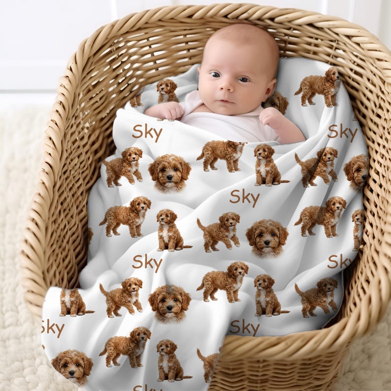 K&ouml;nnte beinhalten: Ein Baby in einem Weidenkorb, eingeh&uuml;llt in eine wei&szlig;e Decke. Die Decke zeigt braune Cavapoo-Welpen-Illustrationen und den Namen "Sky". Die Decke wirkt weich und bequem.