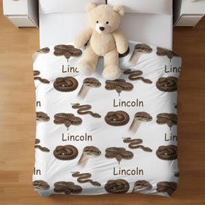 May include: White bedspread with a repeating pattern of brown snakes and the name "Lincoln" in a brown font. A beige teddy bear sits on the bedspread. The bed is made with white sheets and is next to a white nightstand.
