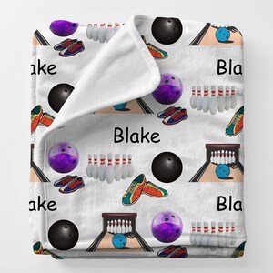 May include: White fleece blanket with a repeating pattern of bowling balls, pins, and shoes in various colors. The word "Blake" is printed in black on the blanket. The blanket is folded over, showing the soft texture.