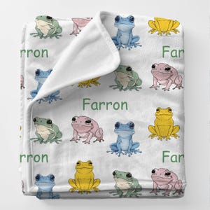 May include: White blanket featuring a repeating pattern of colorful cartoon frogs in shades of green, blue, pink, and yellow. The name "Farron" is printed in green. The blanket is folded.