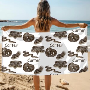 May include: White beach towel with a repeating pattern of brown and grey snakes. The word "Carter" is printed in black on the towel. The towel is being held up on a beach.