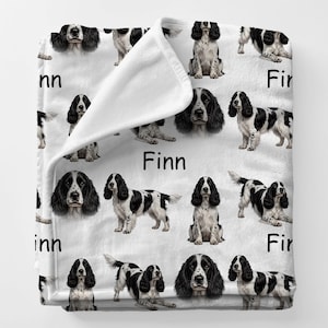 May include: A white blanket featuring a repeating pattern of black and white English Springer Spaniel illustrations. The name "Finn" is printed in black on the blanket.