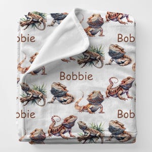 May include: White blanket featuring a repeating pattern of watercolor-style bearded dragons in shades of brown, orange, and green. The blanket also has the names "Bob" and "Bobbie" in brown text.