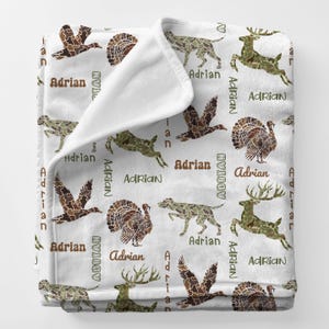 May include: A white fleece blanket featuring a repeating pattern of woodland animals in camouflage colors, including deer, dogs, and turkeys. The name "Adrian" is printed in brown. The blanket is folded over, showing the white underside.