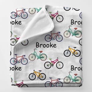 May include: White fleece blanket with a repeating pattern of colorful bicycles in red, teal, yellow, and pink. The name "Brooke" is printed in black on the blanket.