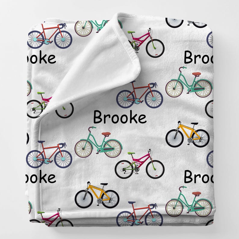 May include: White fleece blanket with a repeating pattern of colorful bicycles in red, teal, yellow, and pink. The name "Brooke" is printed in black on the blanket.