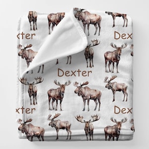May include: White blanket featuring a repeating pattern of brown moose illustrations. The word "Dexter" is printed in brown on the blanket. The blanket is folded over, revealing a soft, plush texture.