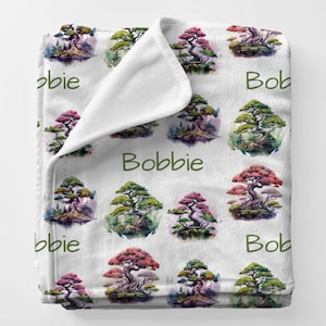 May include: A white plush blanket featuring a repeating pattern of watercolor bonsai trees in shades of green, pink, and red. The name "Bobbie" is printed in green on the blanket. The blanket is folded over, revealing a white interior.
