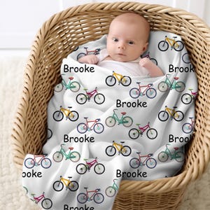 May include: A baby wrapped in a white blanket with colorful bicycle illustrations. The blanket features bicycles in yellow, red, and teal, with the name "Brooke" printed on it. The baby is in a woven basket.