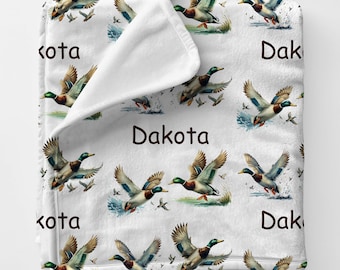 May include: White blanket featuring a repeating pattern of flying ducks in shades of green, brown, and blue. The word "Dakota" is printed in brown. The blanket is folded over, revealing a white interior.