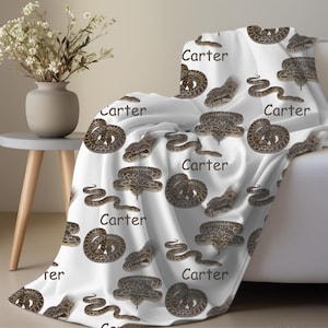 May include: White blanket featuring a repeating pattern of brown snakes and the name "Carter" in black. The snakes are depicted in various poses, some coiled and others slithering. The blanket is draped over a white sofa.