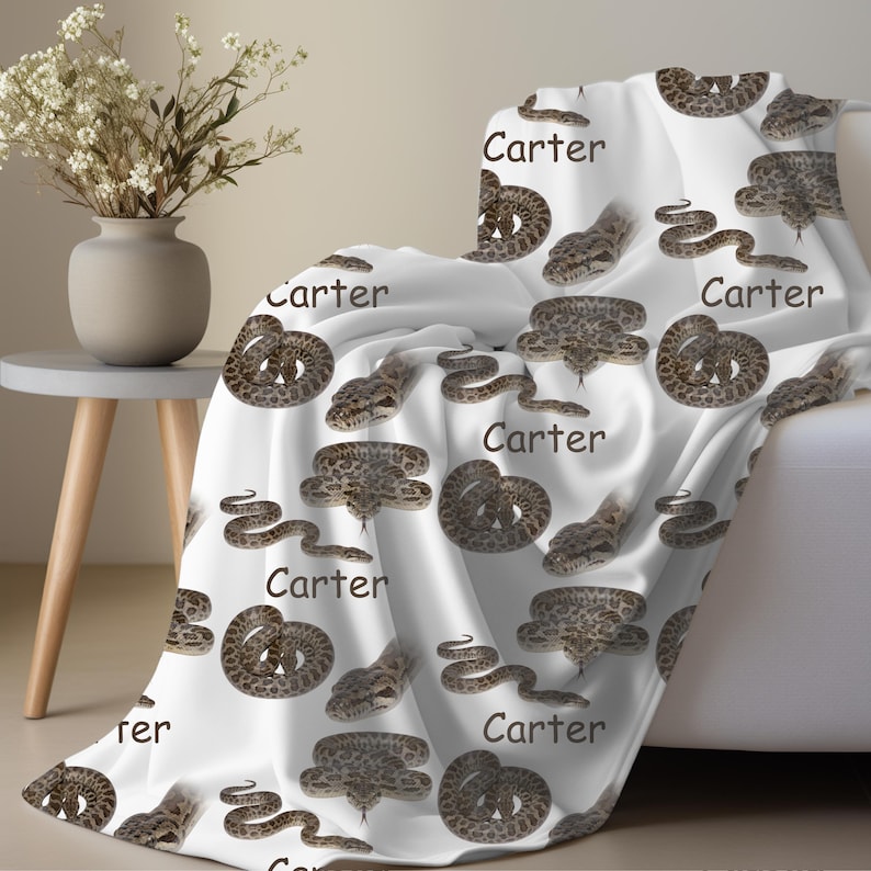 May include: White blanket featuring a repeating pattern of brown snakes and the name "Carter" in black. The snakes are depicted in various poses, some coiled and others slithering. The blanket is draped over a white sofa.