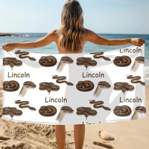May include: White beach towel featuring a repeating pattern of brown snakes and the name "Lincoln" in black. The snakes are in various poses, some coiled and others slithering. The towel is held up on a sandy beach.