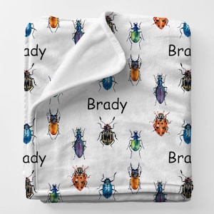 May include: White blanket featuring a repeating pattern of colorful beetles in various shapes and sizes. The name "Brady" is printed in black on the blanket. The blanket is made of a soft, plush material.