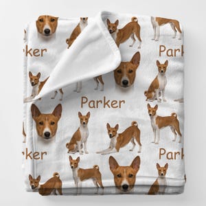 May include: White blanket featuring a repeating pattern of Basenji dogs in various poses. The dogs are brown and white, with the name "Parker" printed in brown. The blanket is folded over, showing the soft texture.