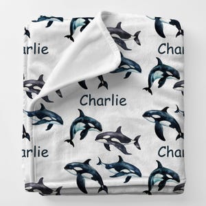 May include: White fleece blanket featuring a pattern of black and white orca whales. The name "Charlie" is printed in dark blue across the blanket. The blanket is folded over, showing the soft, white underside.