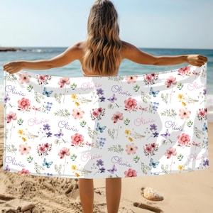 May include: White beach towel with a floral pattern featuring pink, purple, and yellow flowers, blue butterflies, and the name "Olivia" in cursive. The towel is held up on a sunny beach.