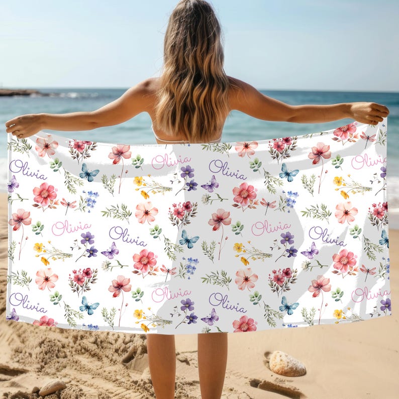 May include: White beach towel with a floral pattern featuring pink, purple, and yellow flowers, blue butterflies, and the name "Olivia" in cursive. The towel is held up on a sunny beach.