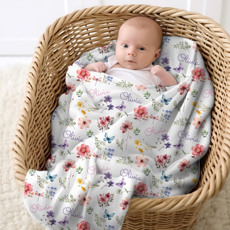 May include: A baby rests in a wicker basket, wrapped in a white floral blanket. The blanket features a pattern of colorful flowers, butterflies, and the name "Olivia" in a script font. The blanket is soft and cozy.