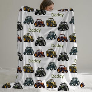 May include: White blanket featuring a repeating pattern of colorful tractors in various shades of green, orange, and yellow. The word "Daddy" is printed in green across the blanket. The blanket is soft and cozy.