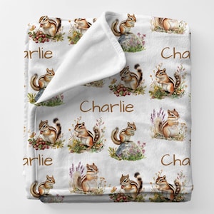 May include: White plush blanket featuring a repeating pattern of watercolor chipmunks, berries, and flowers. The name "Charlie" is printed in brown script across the blanket. The chipmunks are depicted in various poses.