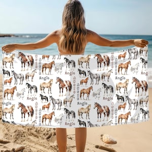May include: A white beach towel featuring a repeating pattern of various horse breeds in shades of brown, black, and gray. The name "Angie" is printed in a vertical column and horizontally across the towel. The towel is being held up on a sandy beach with the ocean in the background.