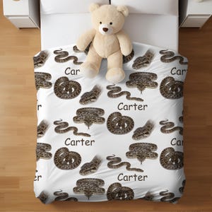 May include: A white bedspread featuring a repeating pattern of brown snakes and the name "Carter" in black. A beige teddy bear rests on the bedspread. The bedspread is on a white bed with white bedside tables either side.