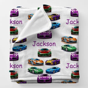 May include: White blanket featuring colorful illustrations of sports cars in various colors, including purple, green, and blue. The name "Jackson" is printed in purple across the blanket.