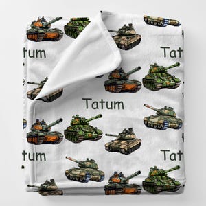 May include: White blanket featuring a repeating pattern of cartoon tanks in various colors, including green, brown, and orange. The name "Tatum" is printed in black. The blanket is folded over, revealing a white interior.
