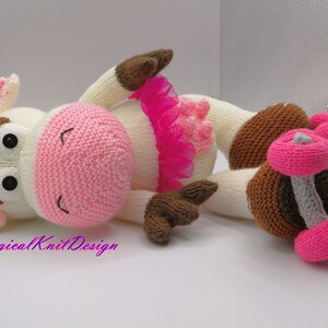 Puede incluir: Una muñeca de vaca de punto blanco con un tutú rosa y patines. La muñeca tiene ojos marrones y una nariz rosa. Los patines son rosas y grises.