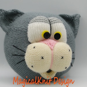 May include: A gray knitted cat head with white and pink facial features, black eyes with yellow pupils, and black whiskers. The cat head is a handmade plush toy.