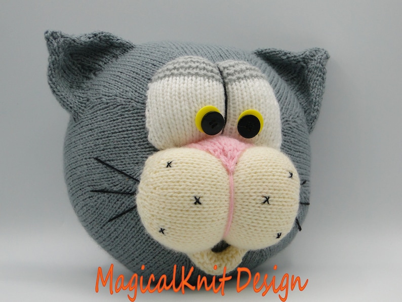 May include: A gray knitted cat head with white and pink facial features, black eyes with yellow pupils, and black whiskers. The cat head is a handmade plush toy.