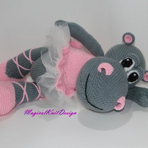 May include: A gray knitted hippopotamus wearing a pink tutu and pink ballet shoes. The hippopotamus is lying on its side with its head resting on its arm. The tutu is made of tulle and has a pink satin ribbon around the waist. The ballet shoes are made of pink felt and have gray laces.