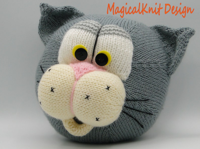 May include: A gray knitted cat head with white and gray stripes, yellow eyes, a pink nose, and black whiskers. The cat head is a knitted toy.