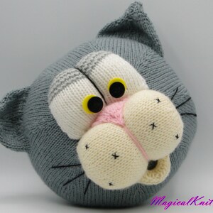 May include: A gray knitted cat head with white and gray stripes on the forehead, yellow eyes, a pink nose, and a white mouth. The cat head is a knitted toy.