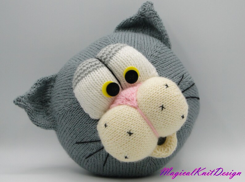 May include: A gray knitted cat head with white and gray stripes on the forehead, yellow eyes, a pink nose, and a white mouth. The cat head is a knitted toy.