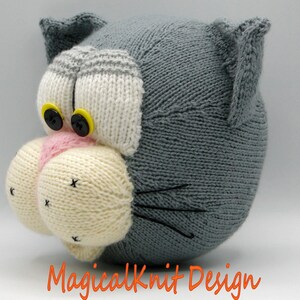 May include: A gray knitted cat head with white and pink details. The cat has black eyes with yellow buttons and black whiskers.