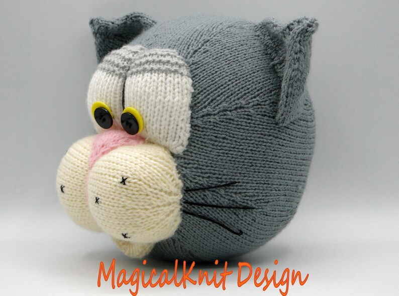 May include: A gray knitted cat head with white and pink details. The cat has black eyes with yellow buttons and black whiskers.