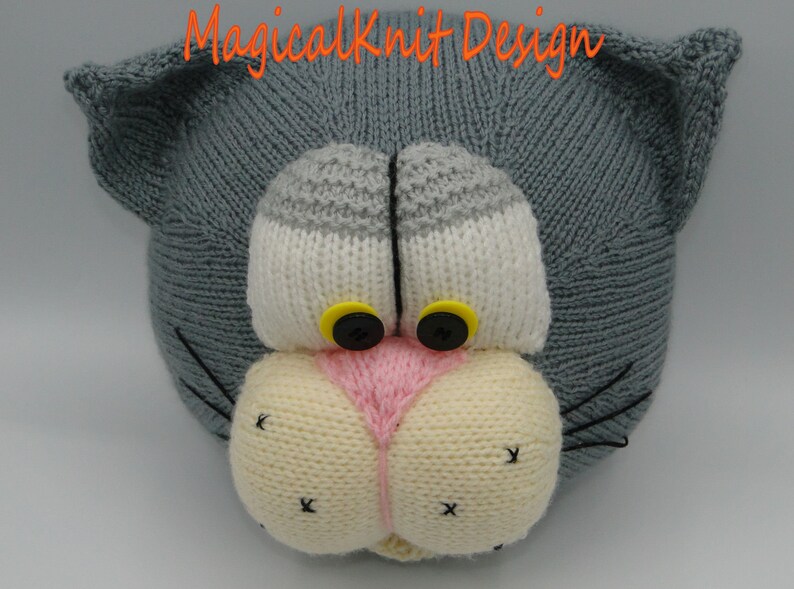 May include: A gray knitted cat head with white and gray eyes, a pink nose, and yellow buttons for eyes. The cat has black whiskers.