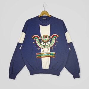 May include: A navy blue sweatshirt with a white and off-white color block design. The sweatshirt features an embroidered design of a stylized geometric pattern with the text "VIA APPIA" in gold lettering.