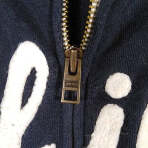 May include: A navy blue zippered hoodie with white embroidered lettering. The zipper is gold metal and has a pull tab.